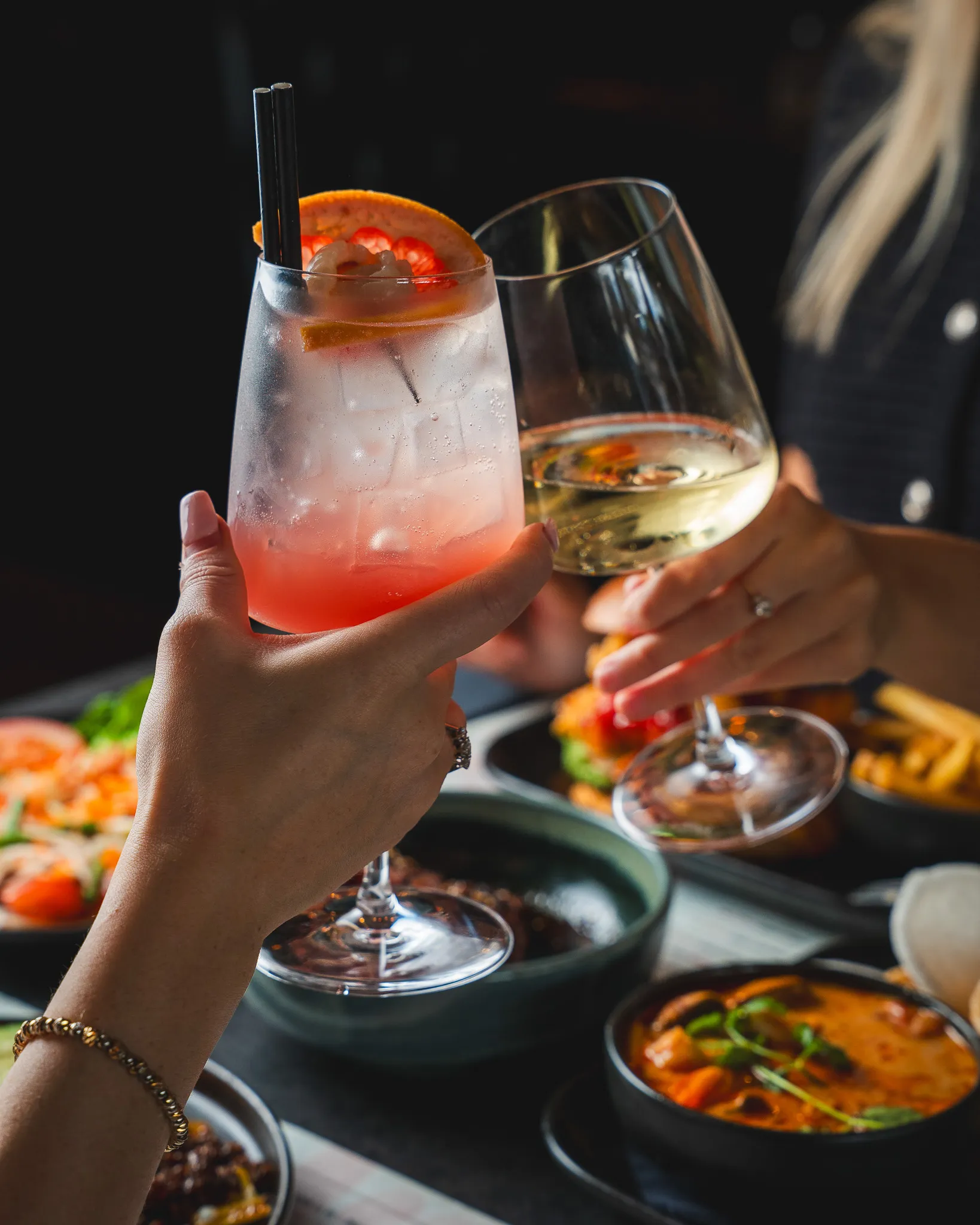 Friends raising a pink cocktail and white wine over dinner.